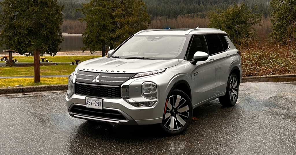 2026-Outlander-PHEV - Humboldt Voice Front-three-quarter view of a silver Mitsubishi Outlander SUV parked on a wet pavement with trees and a lake in the background, daytime outdoor scene.