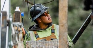 Construction worker in a hard hat, safety vest, and gloves looks up at a perforated metal panel on an outdoor job site.